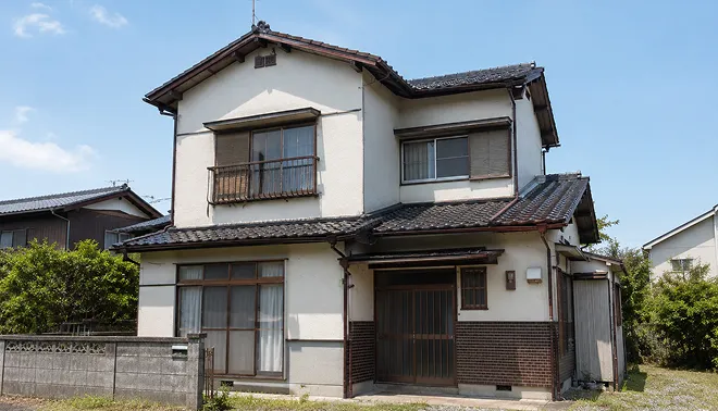手入れが行き届いていない空き家の外観。瓦屋根と白壁の2階建て日本家屋が青空の下に建っている写真