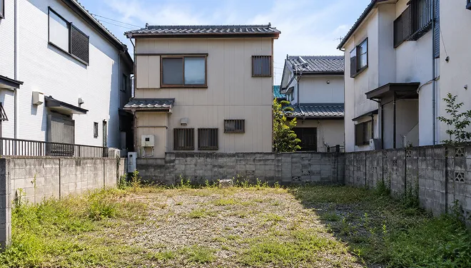 住宅街に位置する空き地。周囲をブロック塀で囲まれ、雑草が生えた更地に隣接する二階建て住宅が並ぶ写真